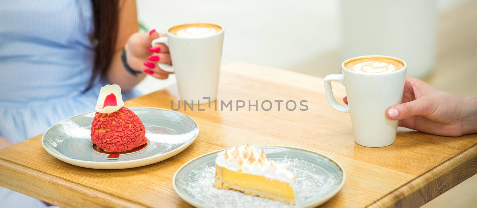 Two young women with cups of coffee and pieces of cake sitting at the table in a cafe outdoors