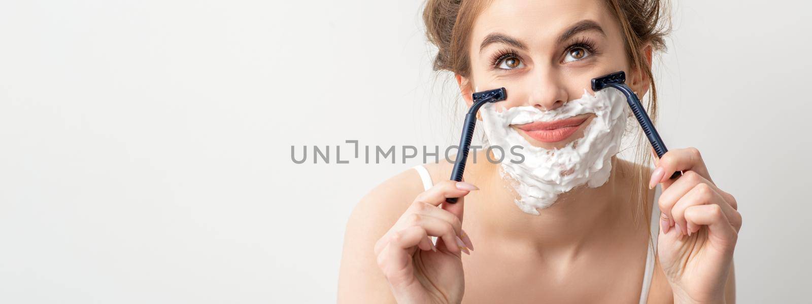 Beautiful young caucasian smiling woman shaving her face with razor looking up on white background