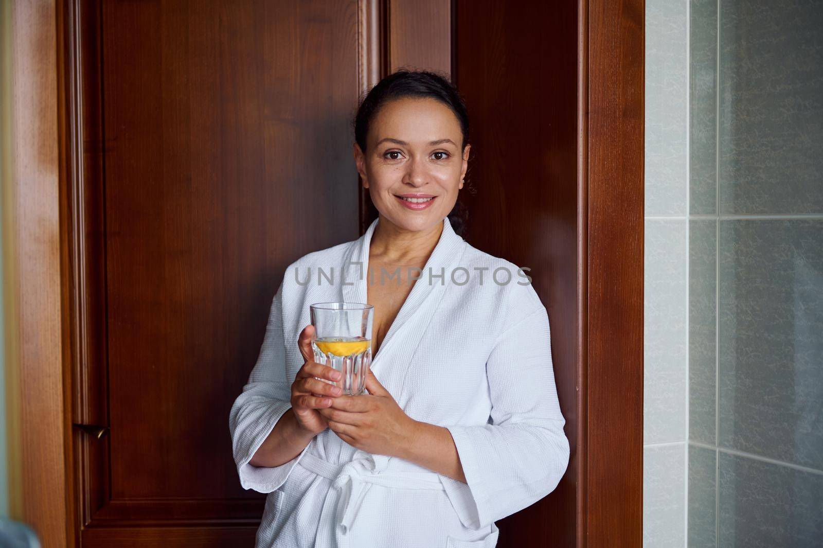 Close-up portrait. Beautiful attractive Hispanic woman with glowing skin, in bathrobe, smiles looking at camera, holds glass with lemon water for good start to the day. Diet, vitality, healthy habits