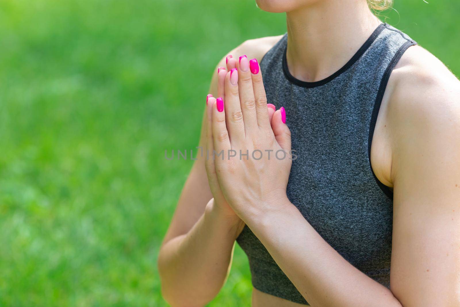 A close-up of some slender woman in a gray top, sitting on the green grass in the park in summer, with her palms folded in front of her. Copy space