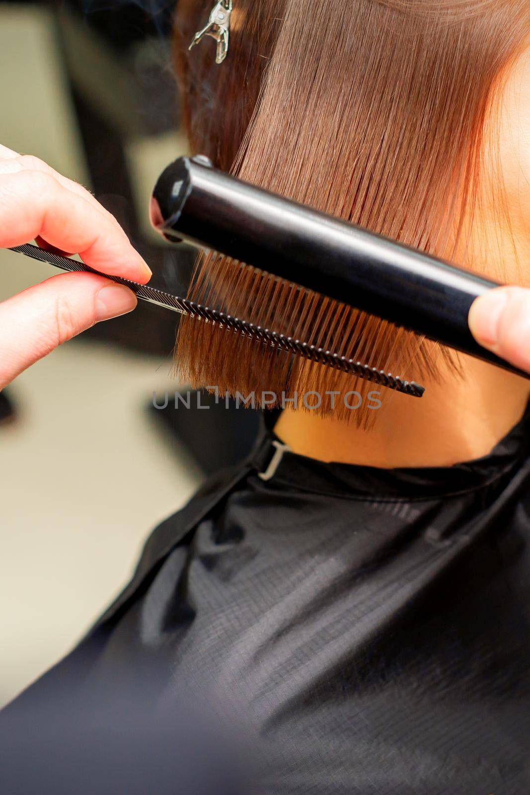 Hair stylist's hands straightening short hair of young brunette woman with flat iron and comb in a beauty salon