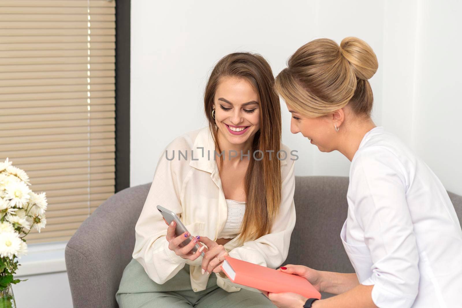 Two young women doctor with pacient smiling and looking at smartphone sitting in hospital office, consultation with a doctor