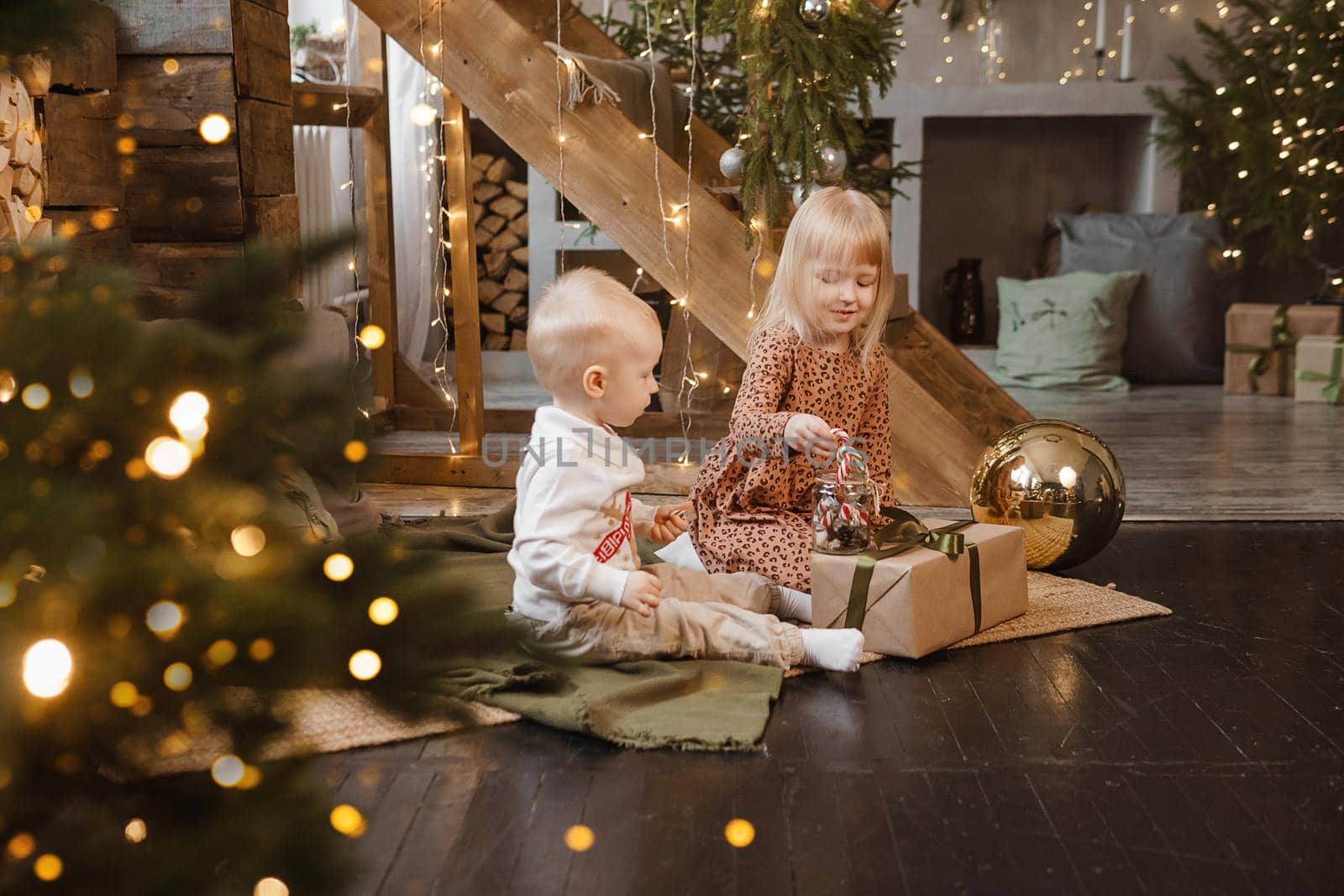 Little brother and sister play on Christmas eve in a beautiful house decorated for the New Year holidays. Children are playing with a Christmas gift. Scandinavian-style interior with live fir trees and a wooden staircase.