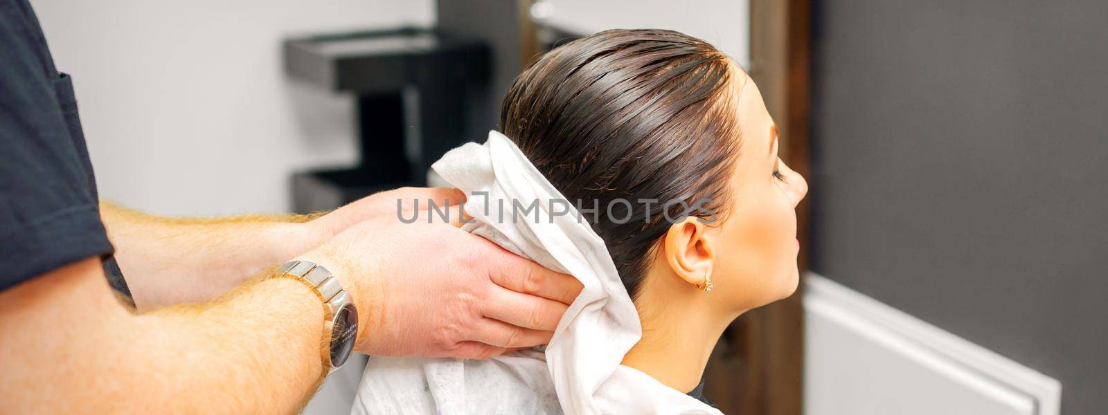 A male hairdresser's hands wipe the hair of a female client with a white towel after shampooing in a beauty salon