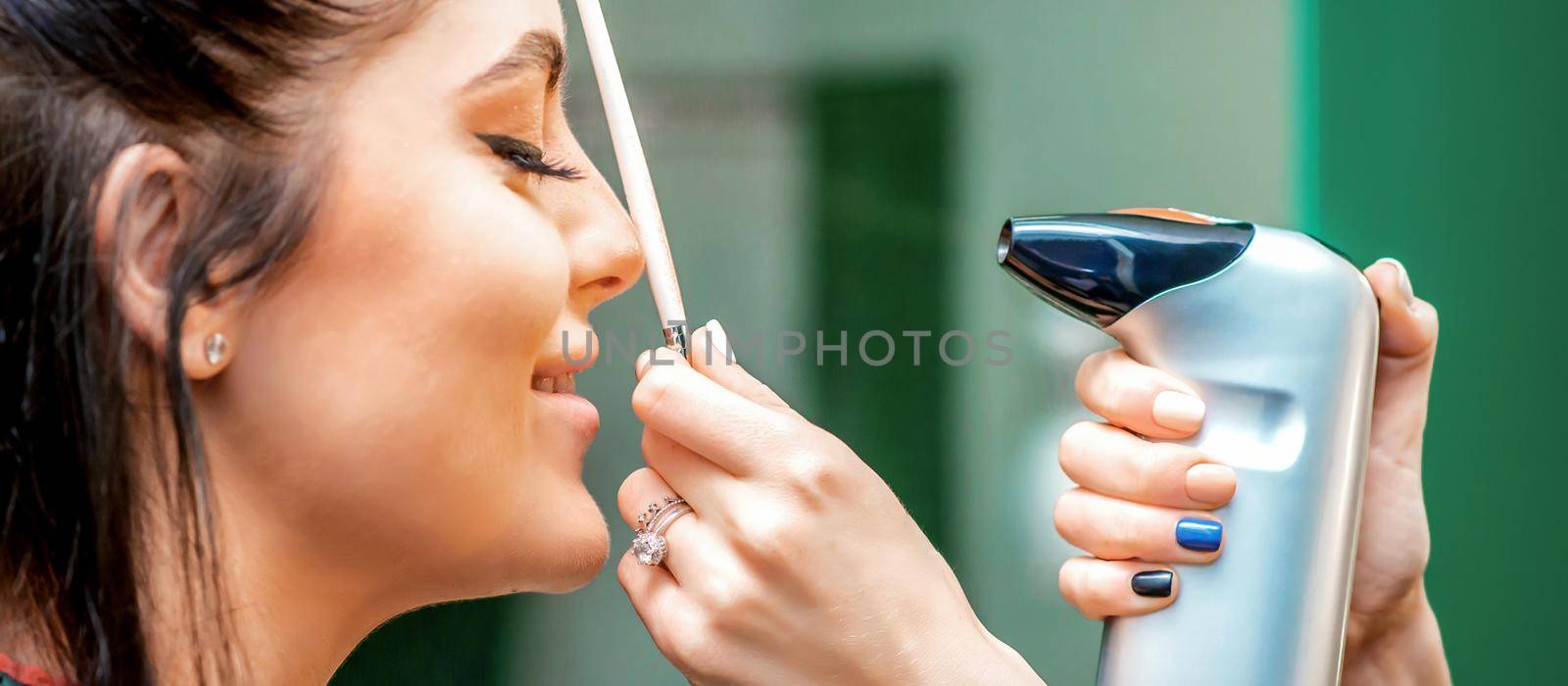 Side view of a make-up artist using aerograph making an airbrush makeup foundation on a female face in a beauty salon
