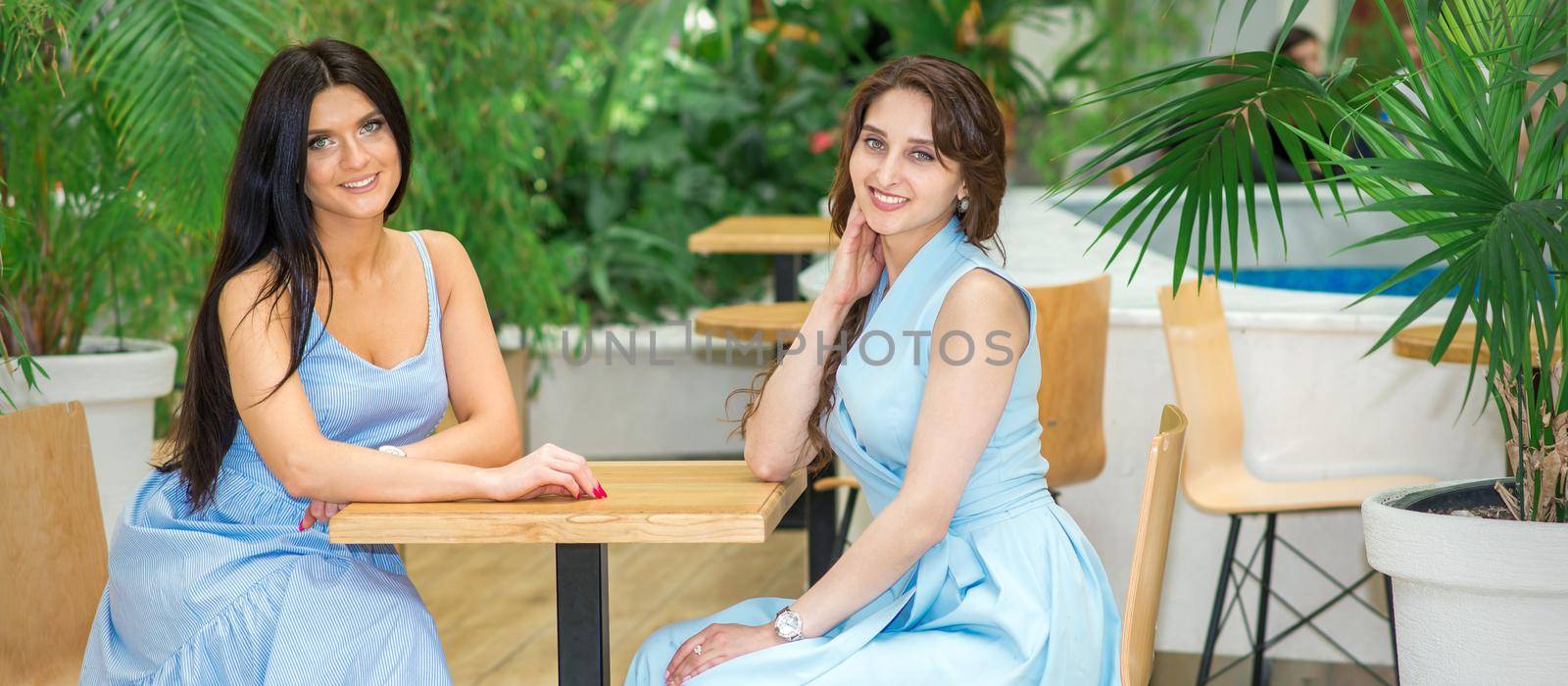 Two beautiful smiling caucasian young women in long blue dresses sitting at the table and looking at the camera in a cafe outdoors