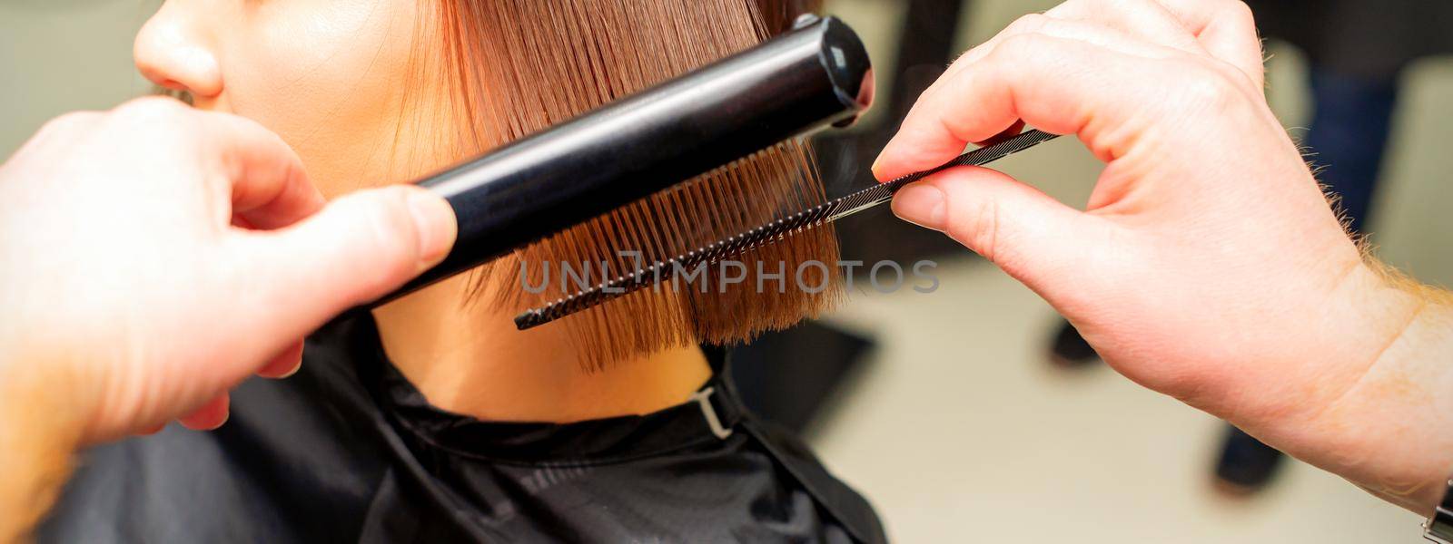 Hair stylist's hands straightening short hair of young brunette woman with flat iron and comb in a beauty salon