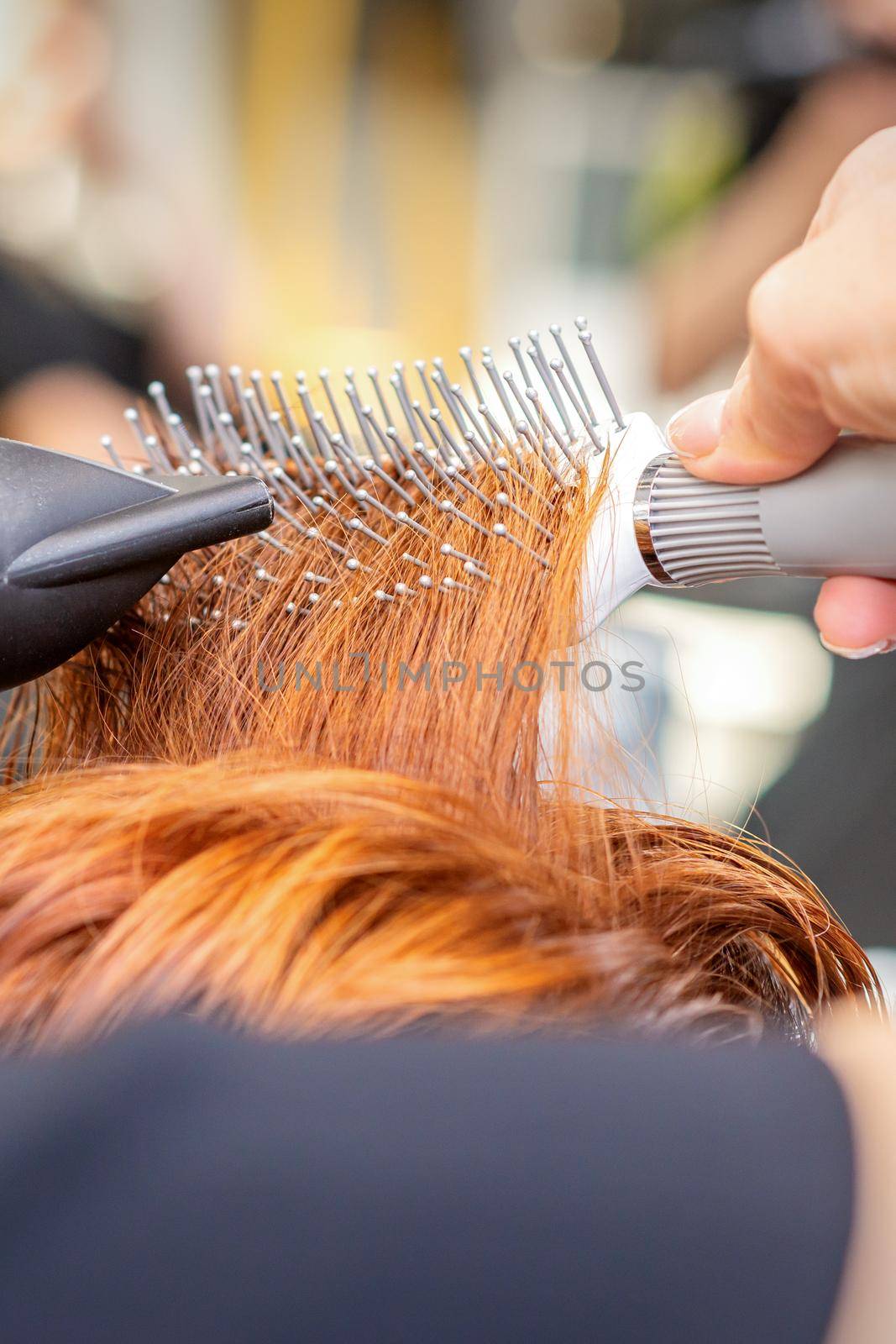 Closeup of master's hand with blow-drying and hairbrush blowing female red hair in a salon