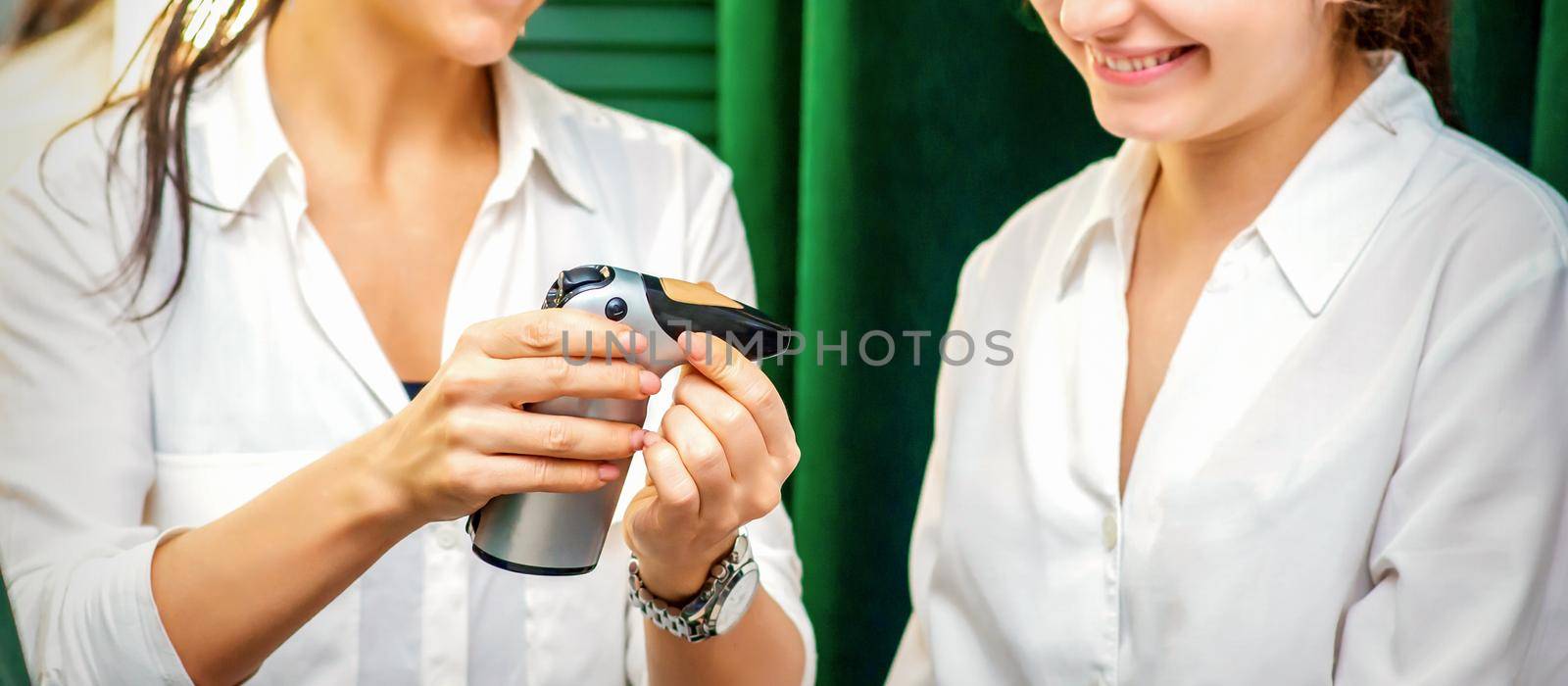 Smiling makeup artist shows for her smiling client aerograph tool device before airbrush procedure in a beauty salon