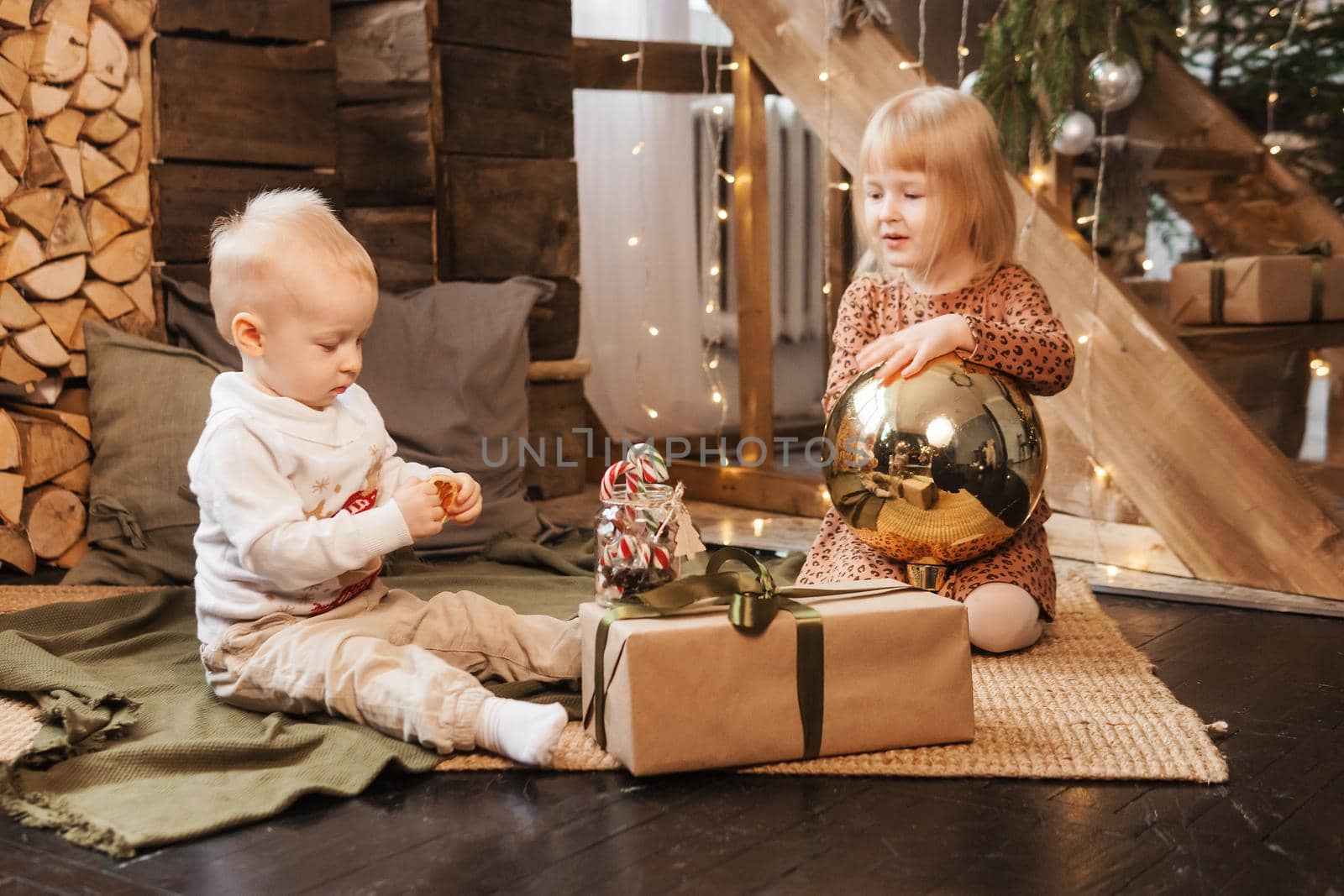 Little brother and sister play on Christmas eve in a beautiful house decorated for the New Year holidays. Children are playing with a Christmas gift. Scandinavian-style interior with live fir trees and a wooden staircase.