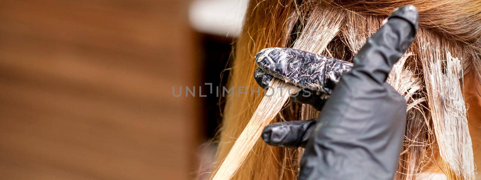 Closeup back view of hairdresser's hands in gloves applying dye to a strand of hair of redhead young woman in a hair salon