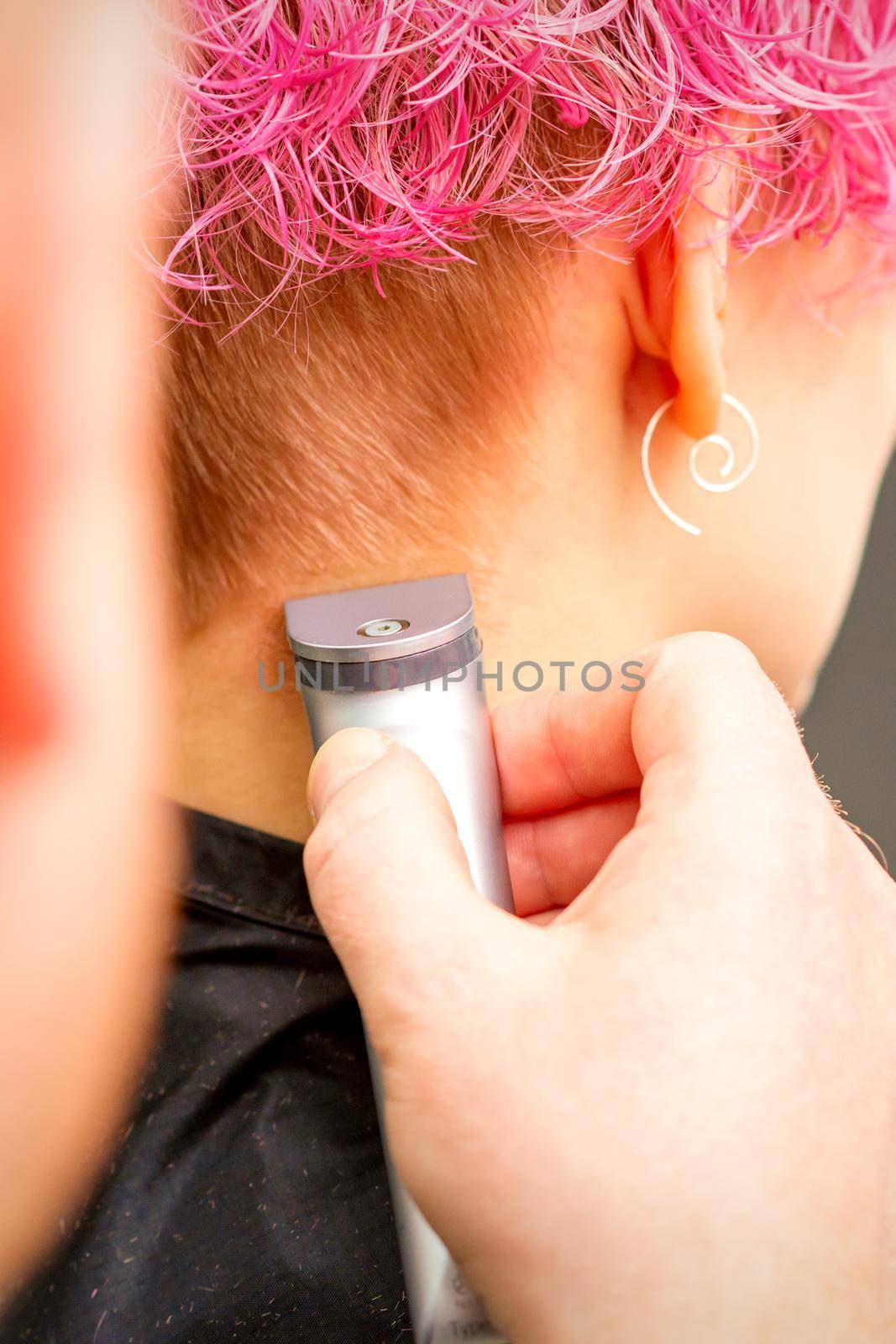 Back view of hairdresser's hand shaving nape and neck with electric trimmer of young caucasian woman with short pink hair in beauty salon