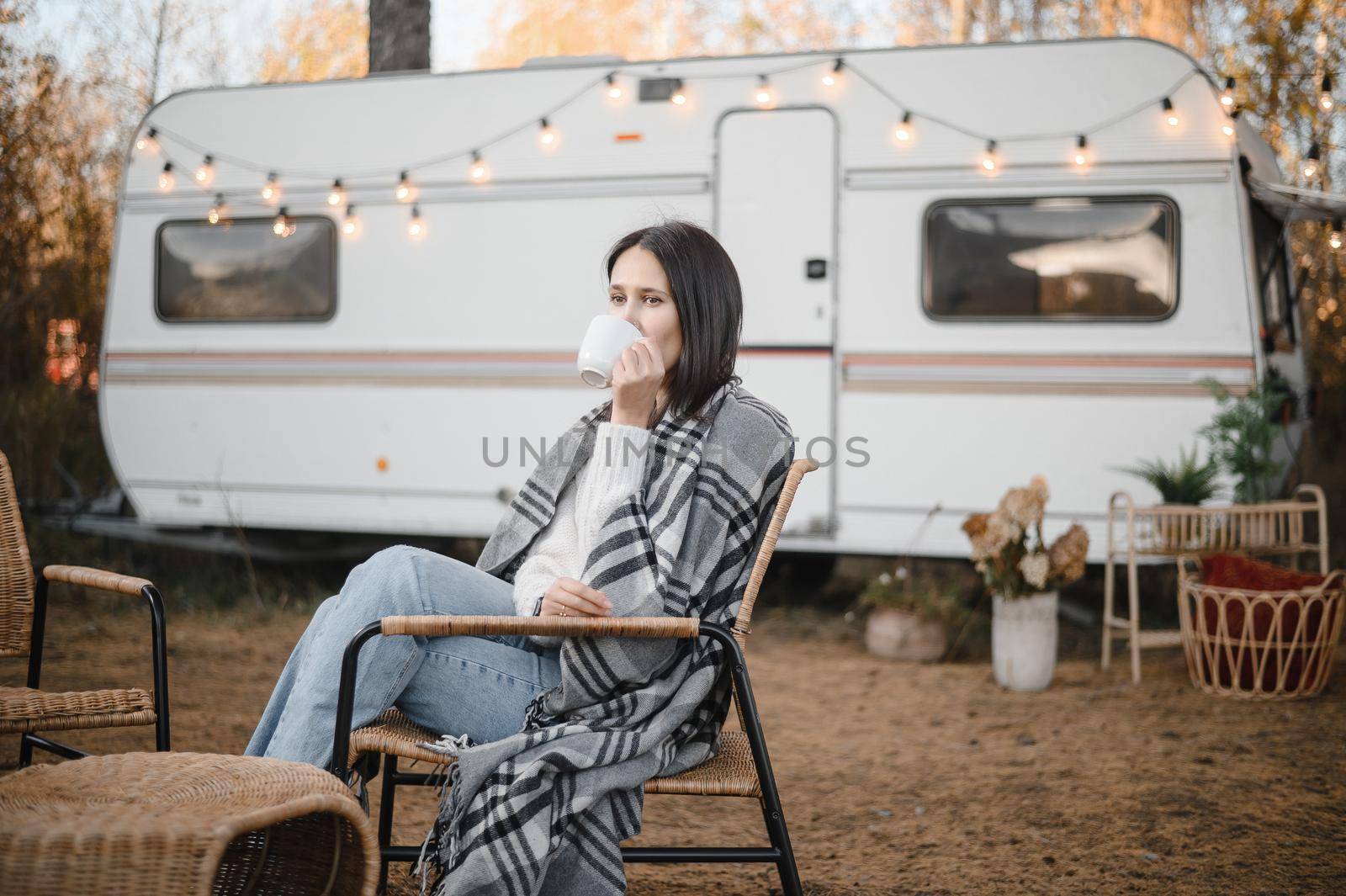 Caucasian woman in a knitted hat wrapped in a plaid and drinks a warming drink outdoors. Travel in a motor home in the fall