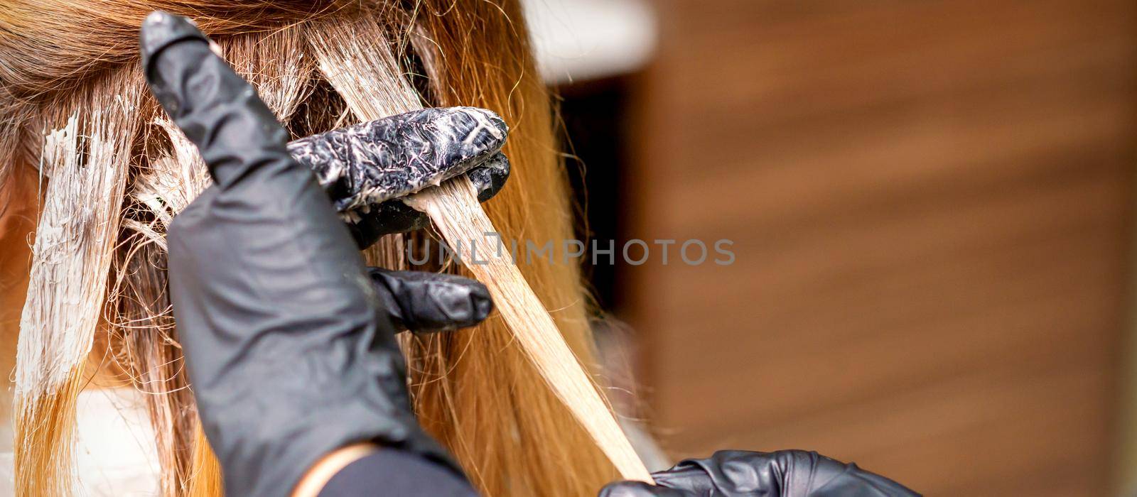 Closeup back view of hairdresser's hands in gloves applying dye to a strand of hair of redhead young woman in a hair salon