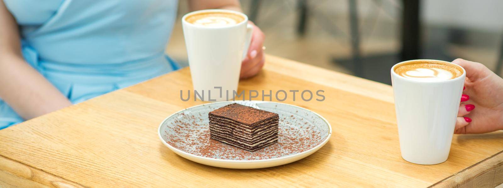 Two young women with cups of coffee and pieces of cake sitting at the table in a cafe outdoors