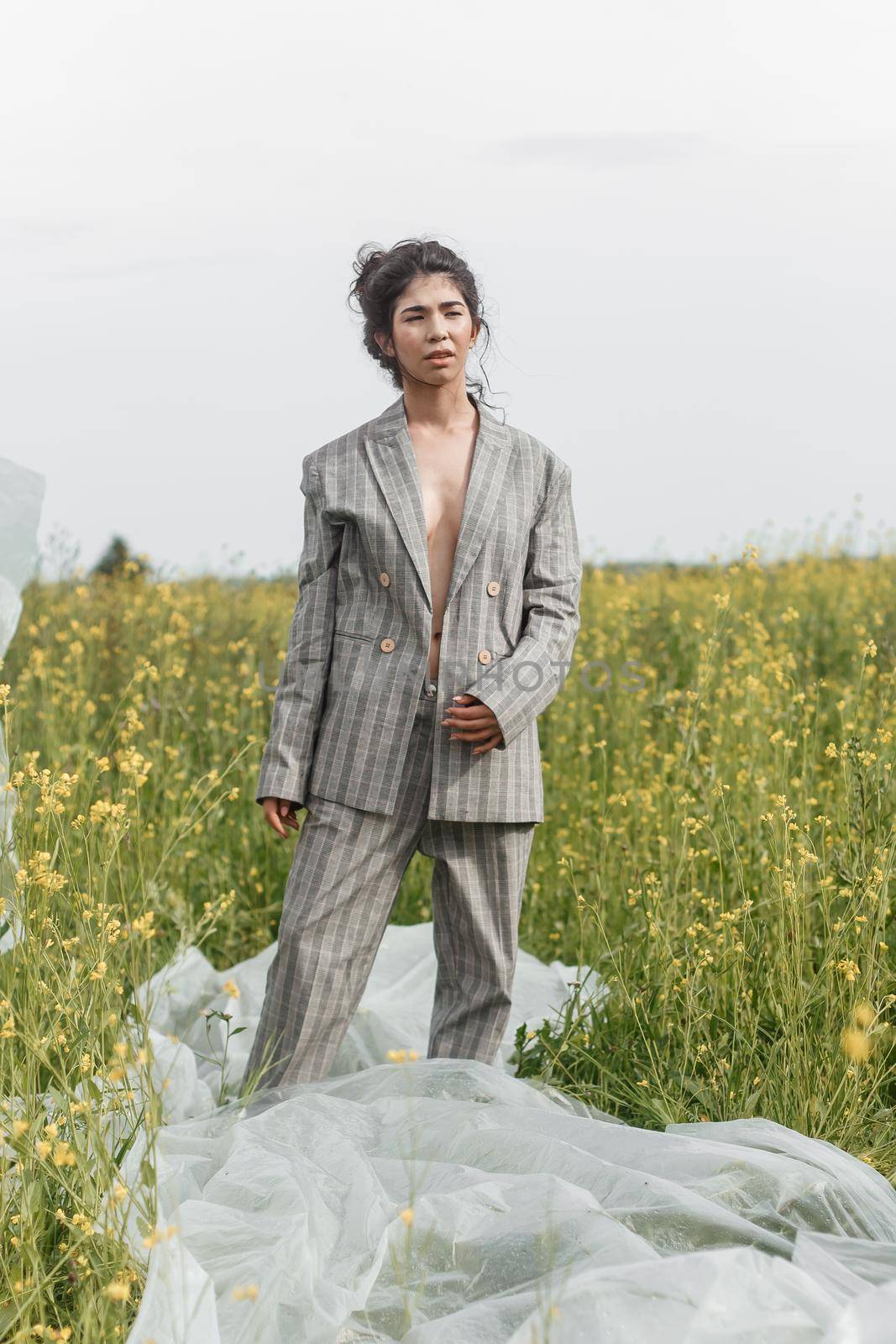 An Asian model poses in a field of yellow flowers for a clothing brand, polyethylene is the main props for a photo shoot. The concept of manufacturing clothing from recycled plastic. A woman in a pantsuit is standing on a plastic bag.