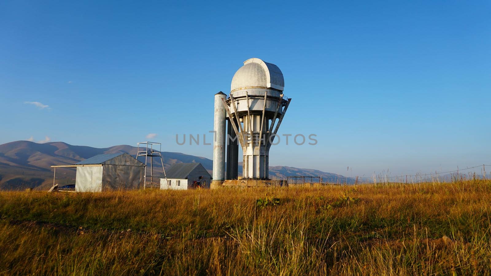Large buildings of the observatory in the form of a dome. The telescopes are closed. Beautiful tall yellow-green grass develops in the wind. Blue sky and green hills in the distance. The Assy Plateau