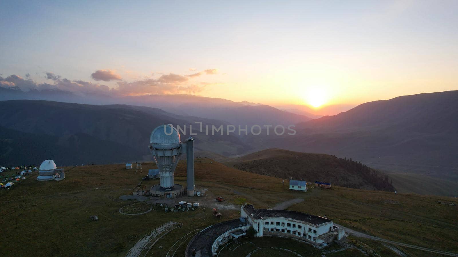 Two large telescope domes at sunset. Drone view of Assy-Turgen Observatory. Beautiful red sunset. Green hills and clouds. Tourists watch the sun. There is a large tent camp and cars nearby. Kazakhstan