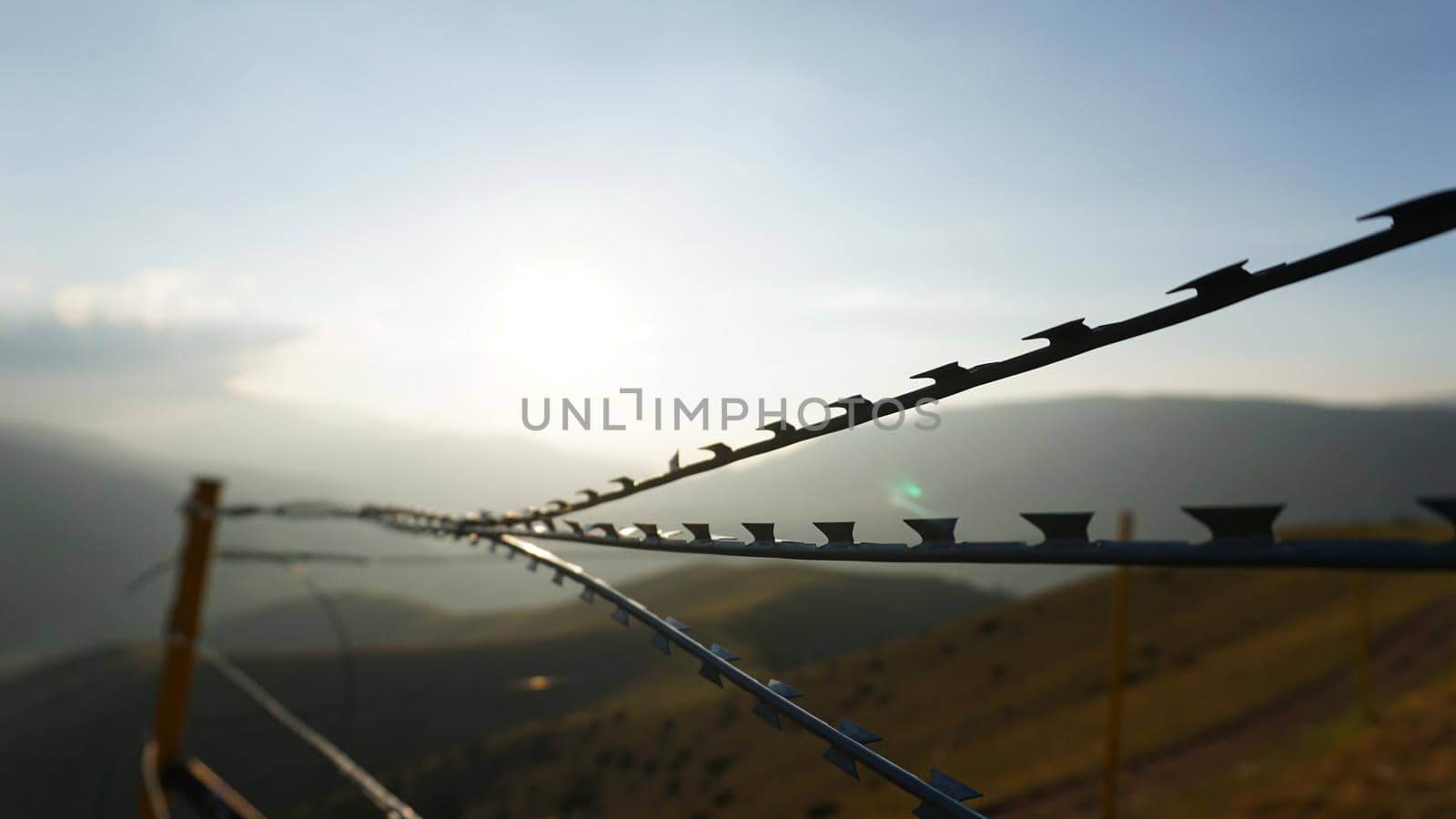 Sharp barbed wire on the fence with a blurred background of mountains. A bright yellow sun in a blue sky. Gray clouds. The high hills are covered with yellow-green grass. Restricted area. Background