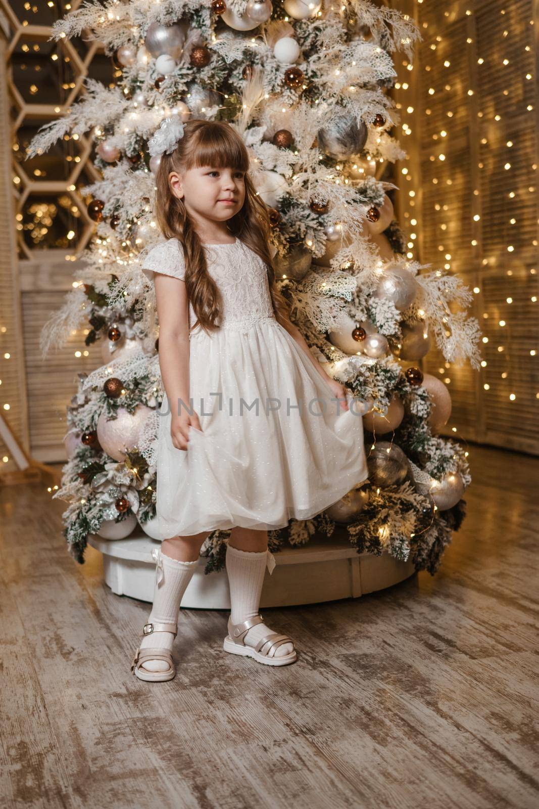 A girl in a festive light dress with a gift in her hands next to a Christmas tree, lights of garlands in the background. The concept of New Year holidays.