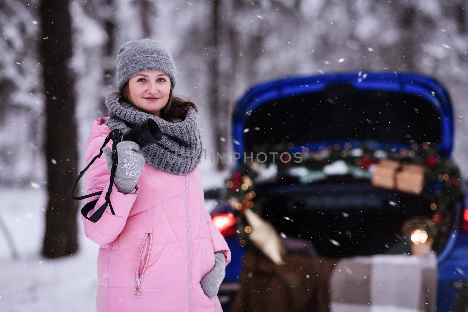 A woman in a winter snow-covered forest in the trunk of a car decorated with Christmas decor. A female photographer holds a camera in her hands.