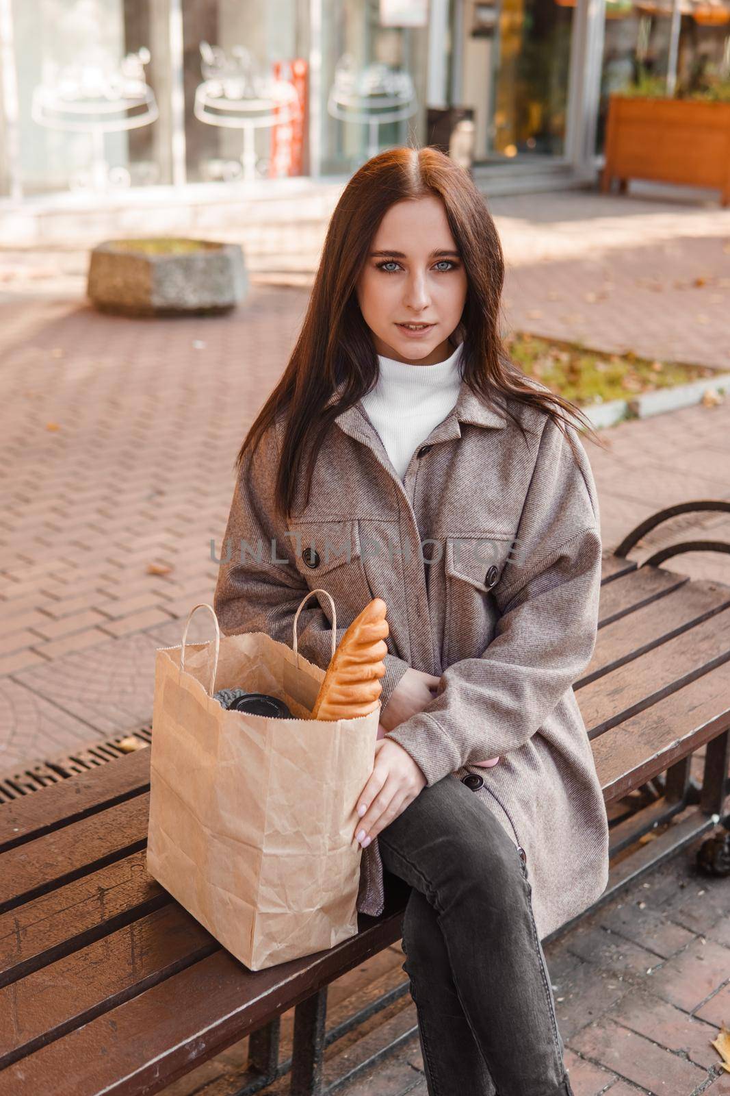 A stylish brunette woman walks around the autumn city. The brunette is sitting on a bench outside with a glass of coffee and a phone, holding a craft bag with a baguette.