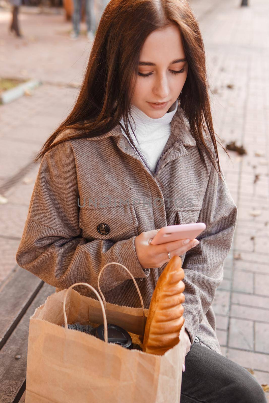 A stylish brunette woman walks around the autumn city. The brunette is sitting on a bench outside with a glass of coffee and a phone, holding a craft bag with a baguette.