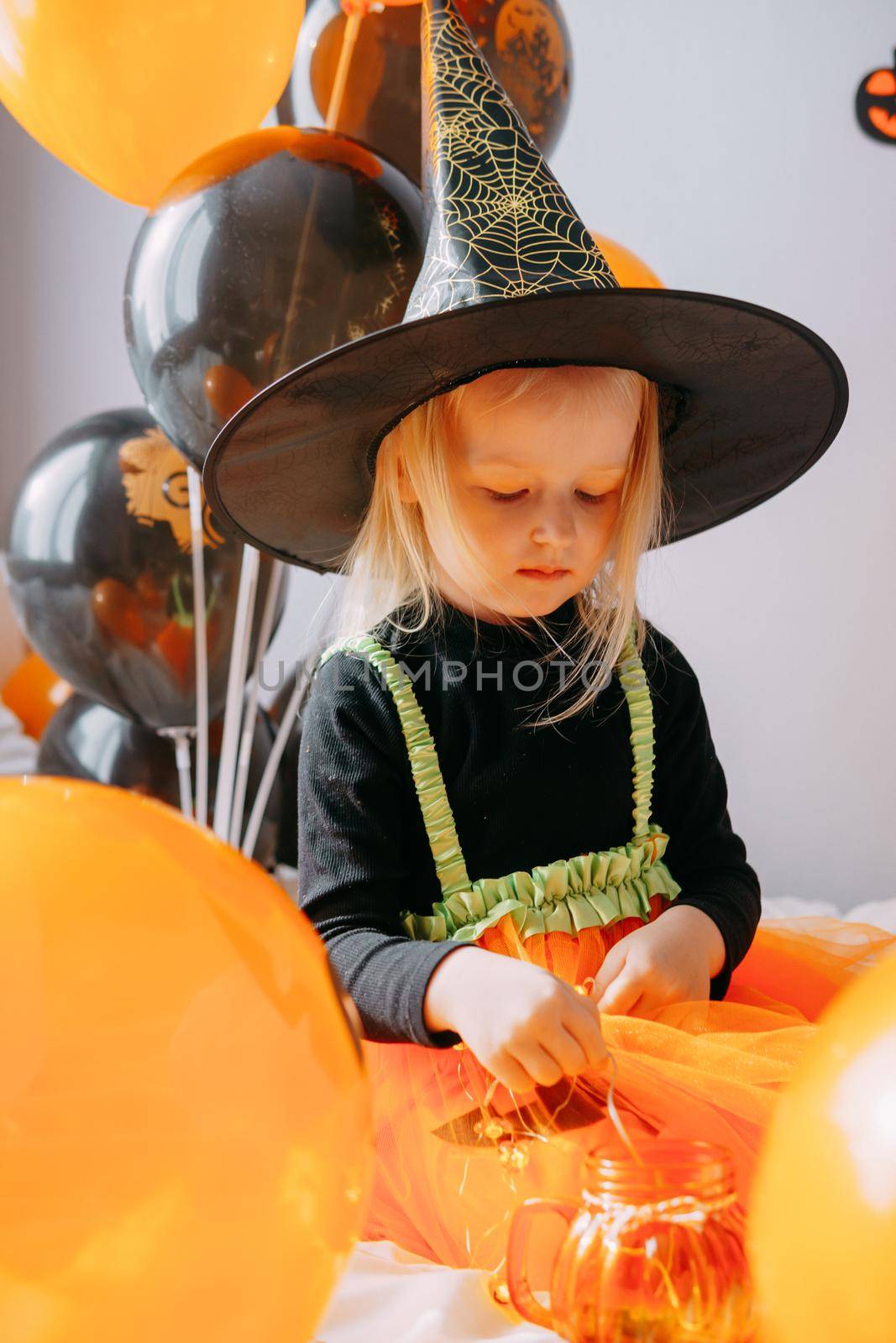 Children's Halloween - a girl in a witch hat and a carnival costume with airy orange and black balloons at home. Ready to celebrate Halloween.