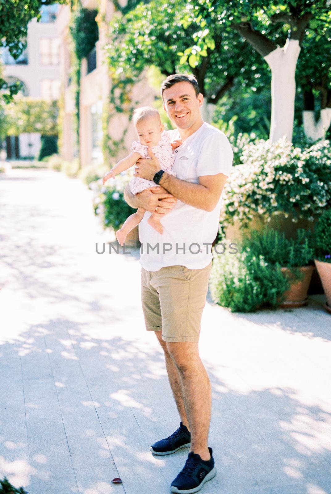 Smiling dad with a little girl in his arms stands on the street near flower pots by Nadtochiy
