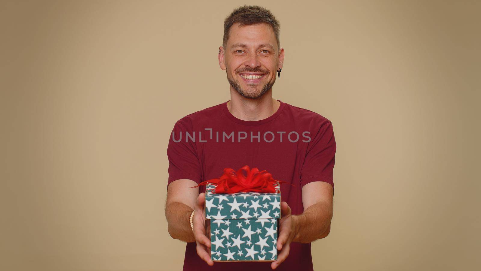 Positive smiling bearded handsome young man presenting birthday gift box stretches out hands, offer wrapped present career bonus, celebrating party. Adult stylish guy on beige studio background indoor