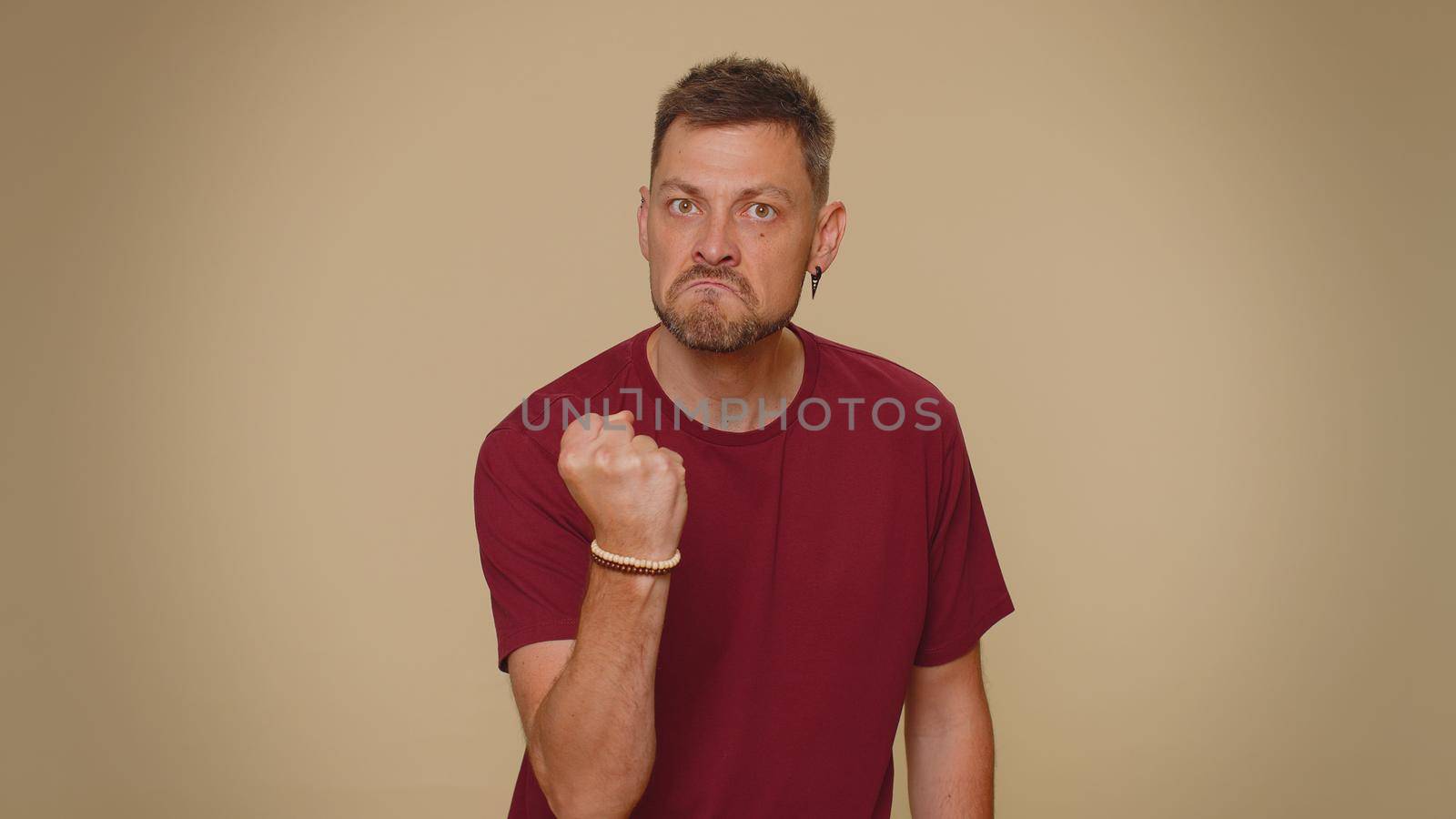 Aggressive angry handsome young man in red t-shirt trying to fight at camera, shaking fist, boxing with expression, punishment. Adult stylish male guy isolated alone on beige studio background indoors