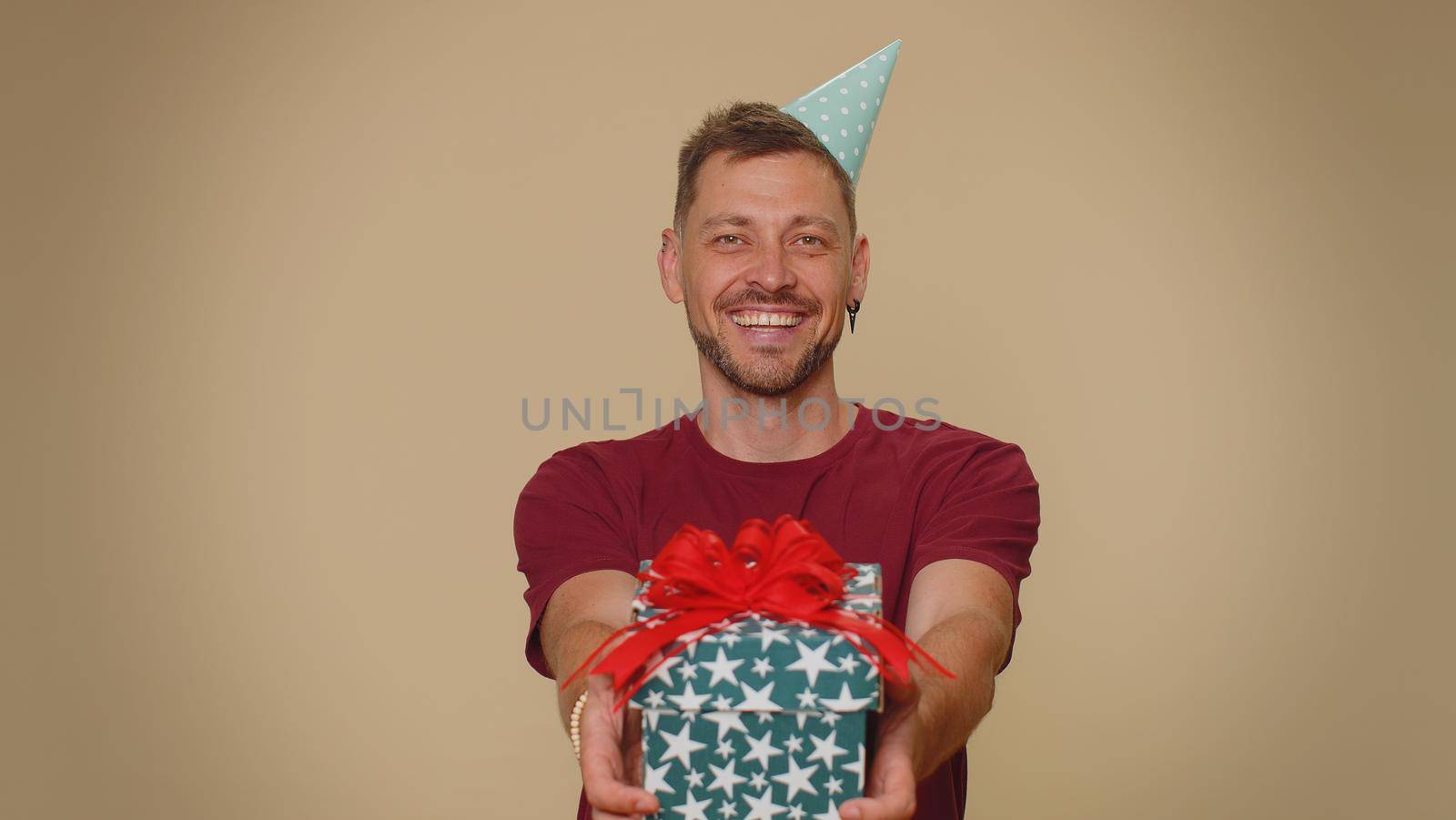 Positive smiling bearded handsome young man presenting birthday gift box stretches out hands, offer wrapped present career bonus, celebrating party. Adult stylish guy on beige studio background indoor