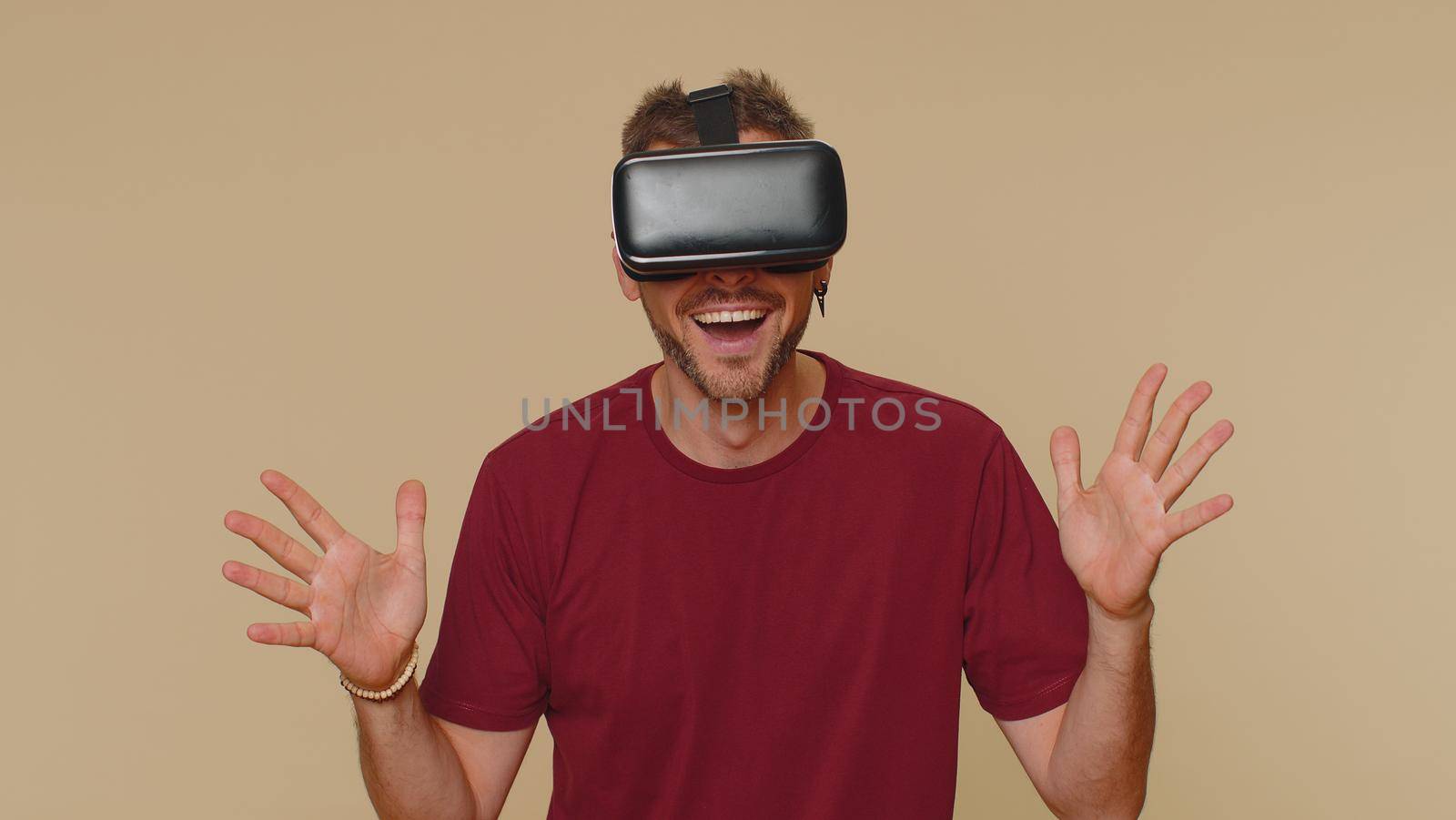 Bearded handsome young man using headset helmet app to play simulation drawing game. Watching virtual reality 3D 360 video. Adult male guy in VR goggles isolated on beige studio background indoors