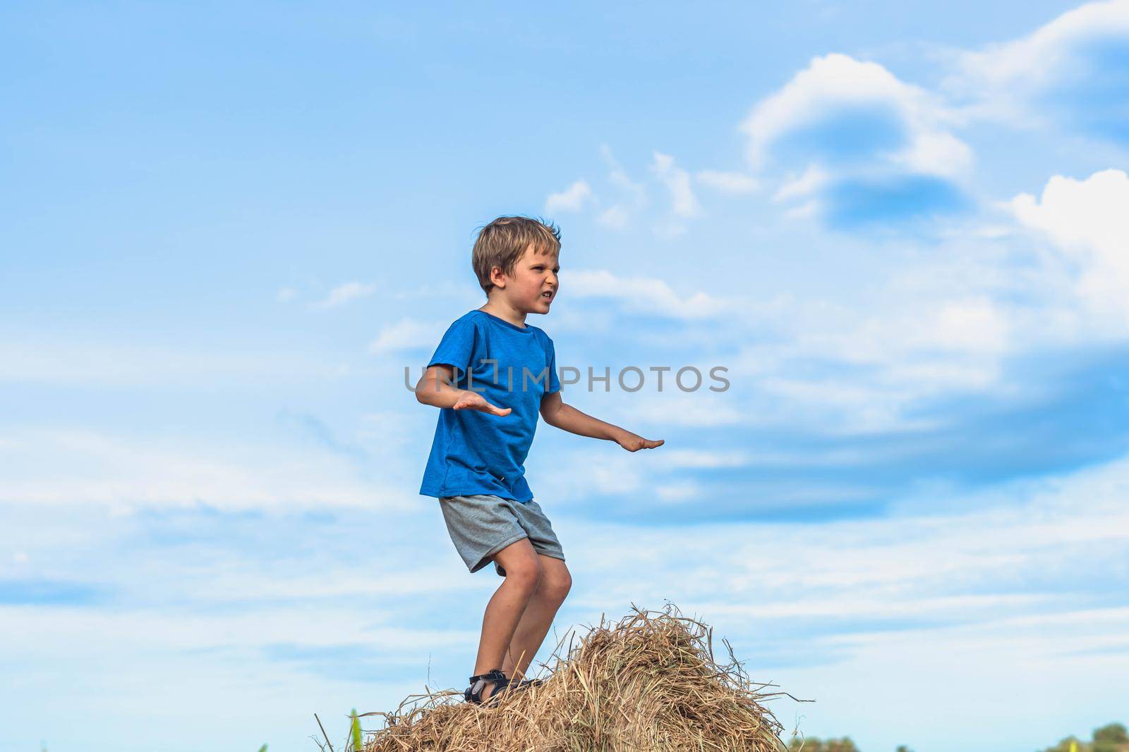 Boy smile play dance grimace show off blue t-shirt stand on haystack bales of dry grass, clear sky sunny day. Balance training. Concept happy childhood, children outdoors, clean air close to nature.