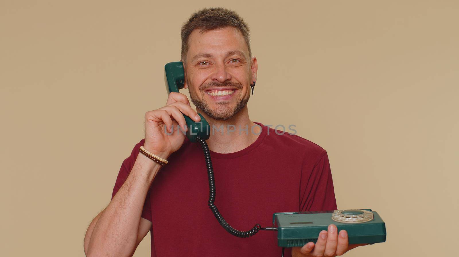 Hey you, call me back. Handsome young man in red t-shirt talking on wired vintage telephone of 80s, says hey you call me back. Call center. Adult stylish male guy on beige studio background indoor