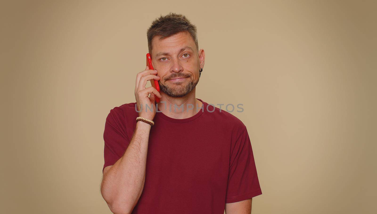 Tired young man in red t-shirt making online conversation speaks on smartphone. Boring call. Blah expression. Nonsense talking. Adult stylish male guy isolated alone on beige studio wall background