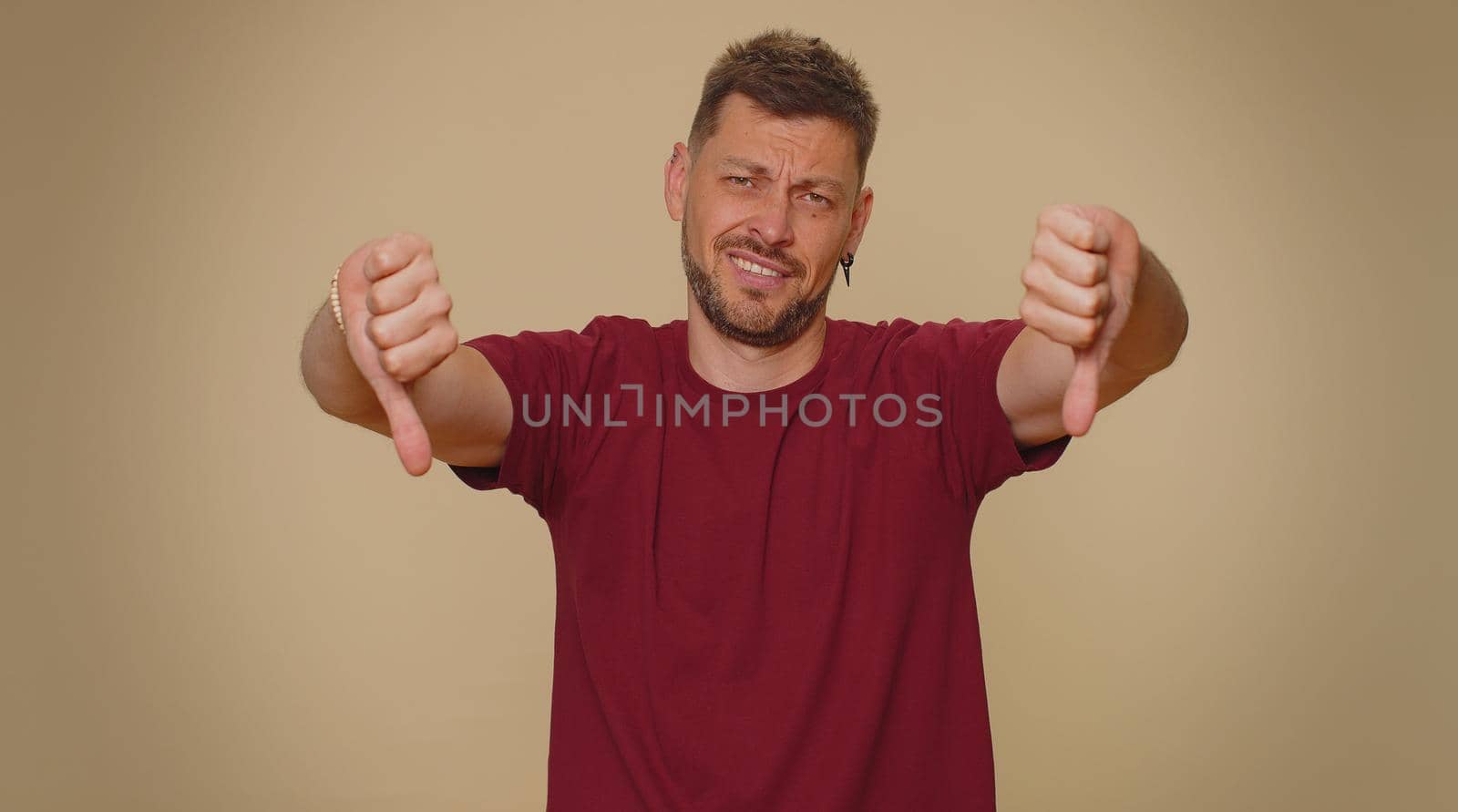 Upset bearded handsome young man showing thumbs down sign gesture, expressing discontent, disapproval, dissatisfied, dislike. Adult stylish male guy isolated alone on beige studio background indoors