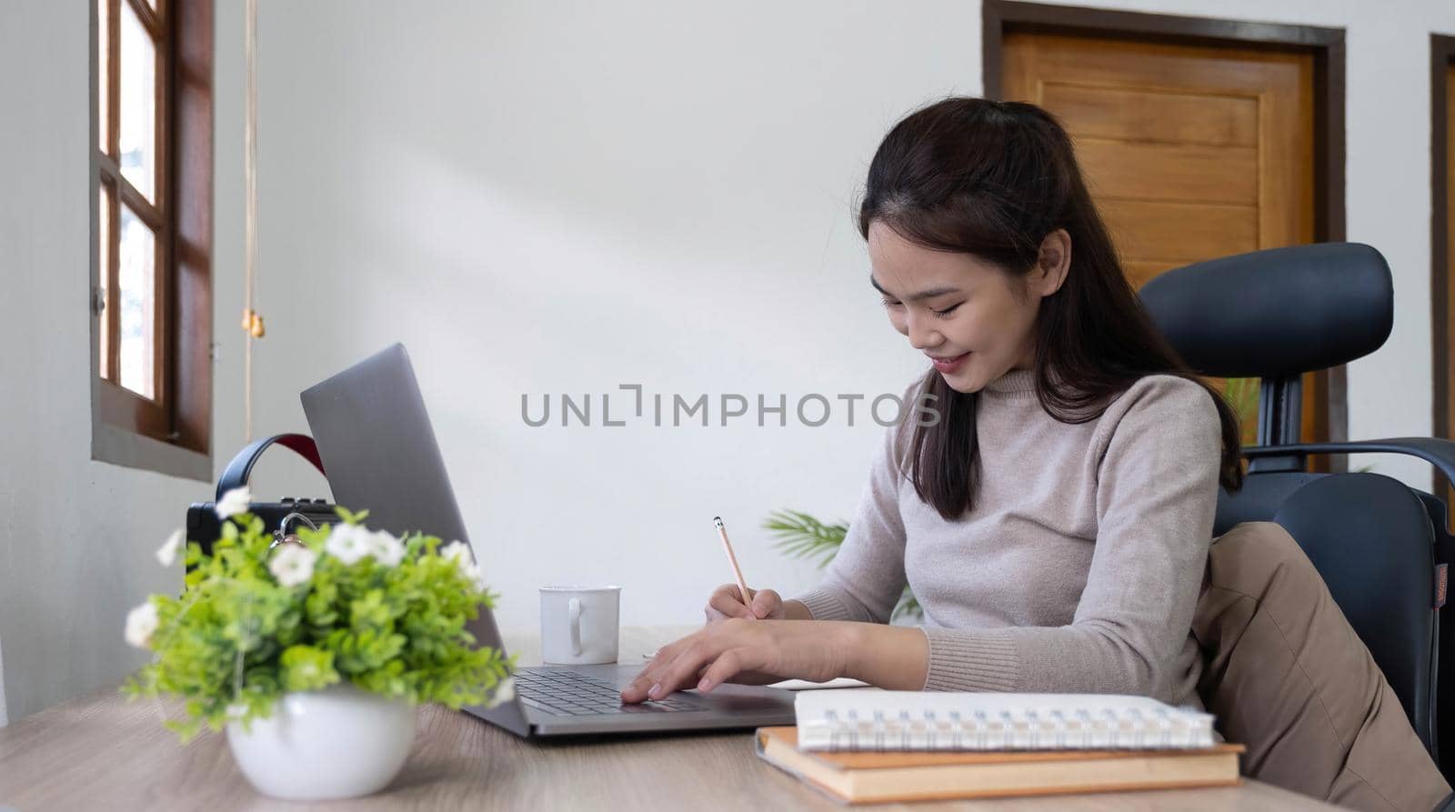Beautiful woman writing taking notes while sitting in front her computer laptop at the wooden working table over living room bookshelf as background