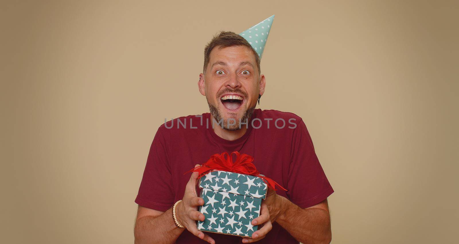 Cheerful bearded handsome young man receiving gift box and smiling joyfully, looking amazed satisfied with nice present, unexpected birthday surprise. Stylish male guy on beige studio background
