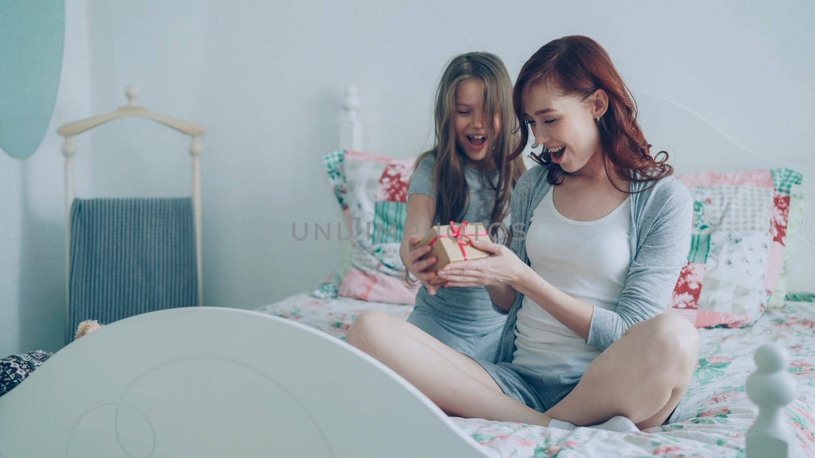 Little cute girl giving gift box to her young happy mother celebrating birthday sitting on bed in bright bedroom at home