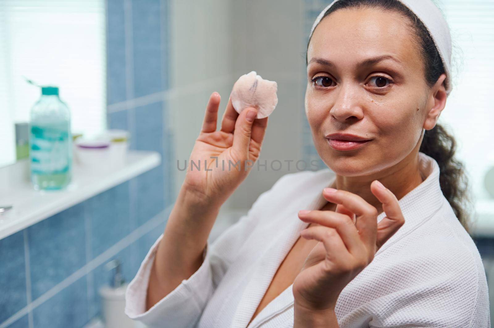 Healthy skin is a daily commitment. Close-up portrait of a charming middle-aged woman in white bathroom removing make-up using, showing a cotton pad while looking confidently at the camera. Skin care