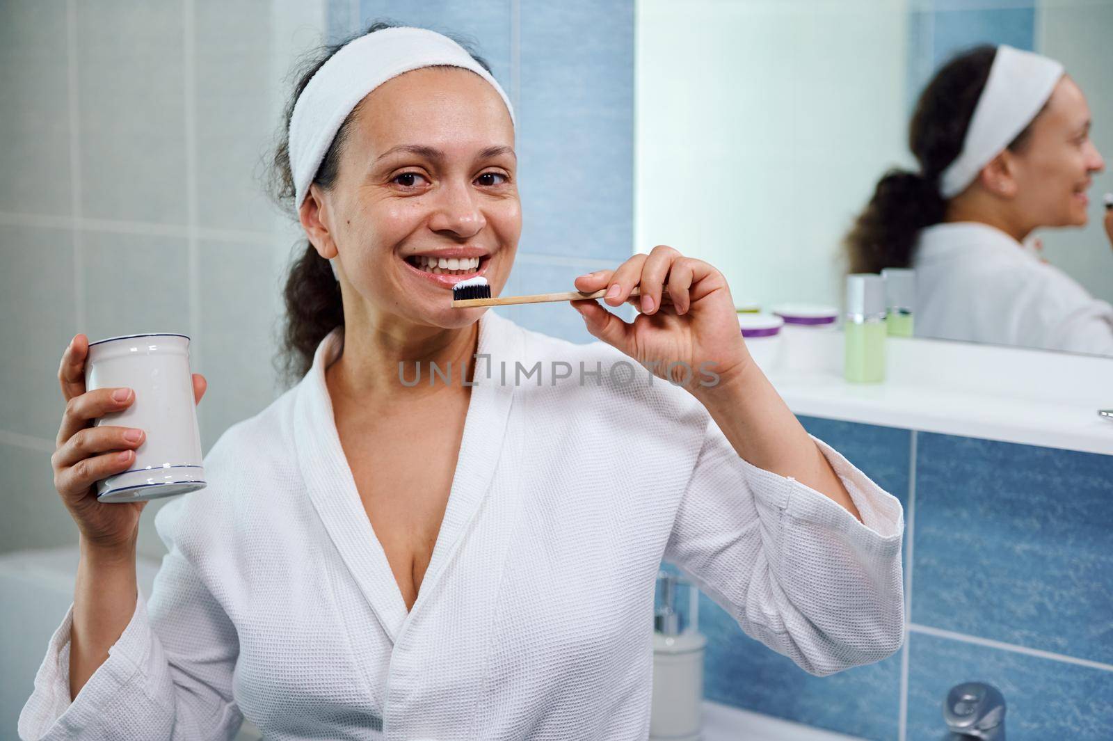 Happy Latin American woman in white bathrobe, brushing teeth with plastic free toothbrush and whitening toothpaste smiling beautiful toothy smile while looking at camera. Oral hygiene and dental care