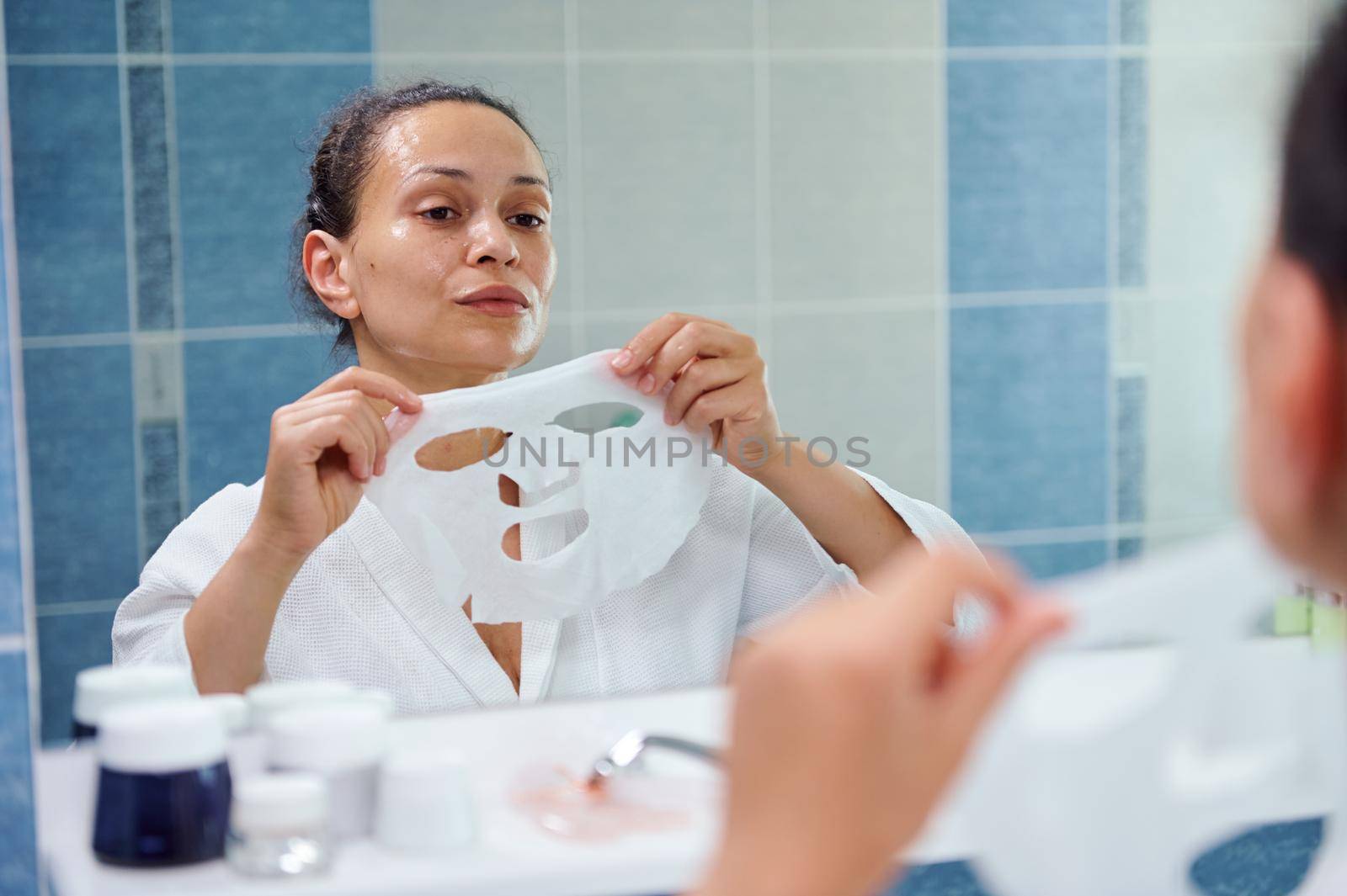 Reflection in bathroom mirror of a beautiful dark-haired multi-ethnic woman, in white bathrobe applying a smoothing moisturizing textile face mask. Skin care. Home spa. Anti-aging beauty procedures