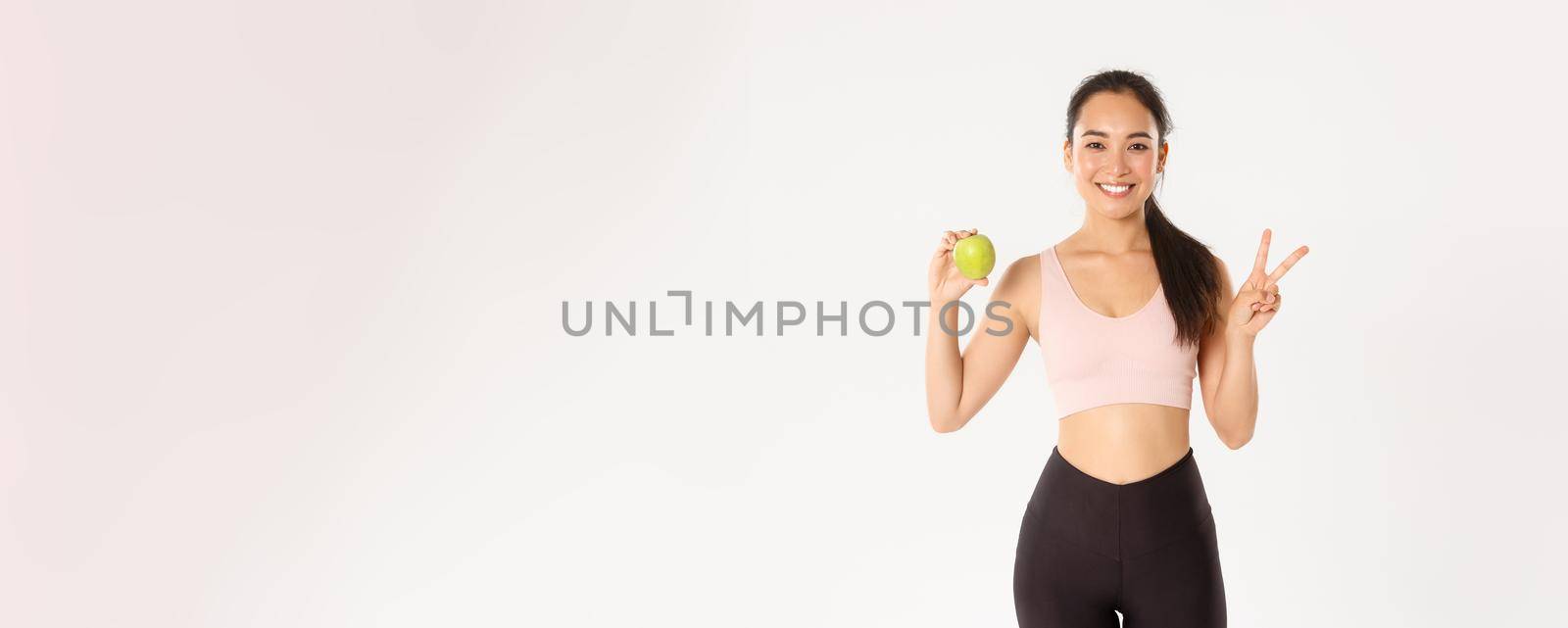 Sport, wellbeing and active lifestyle concept. Attractive asian female fitness coach, girl trainer advice eating healthy food after workout and training, standing with apple, show peace sign by Benzoix