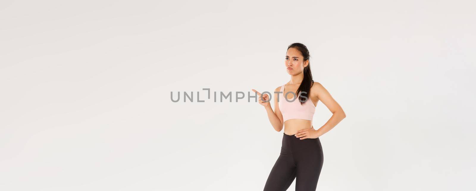 Sport, gym and healthy body concept. Full length of skeptical and displeased female athlete, asian sportswoman sulking and frowning disappointed, pointing finger upper left corner and complaining.