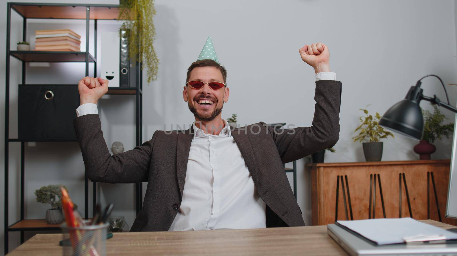 Businessman wears sunglasses, festive cap, listening music at workplace dancing victory dance, weekends. Rejoicing developing project. Celebrating success. Lottery jackpot. Motivated people in office