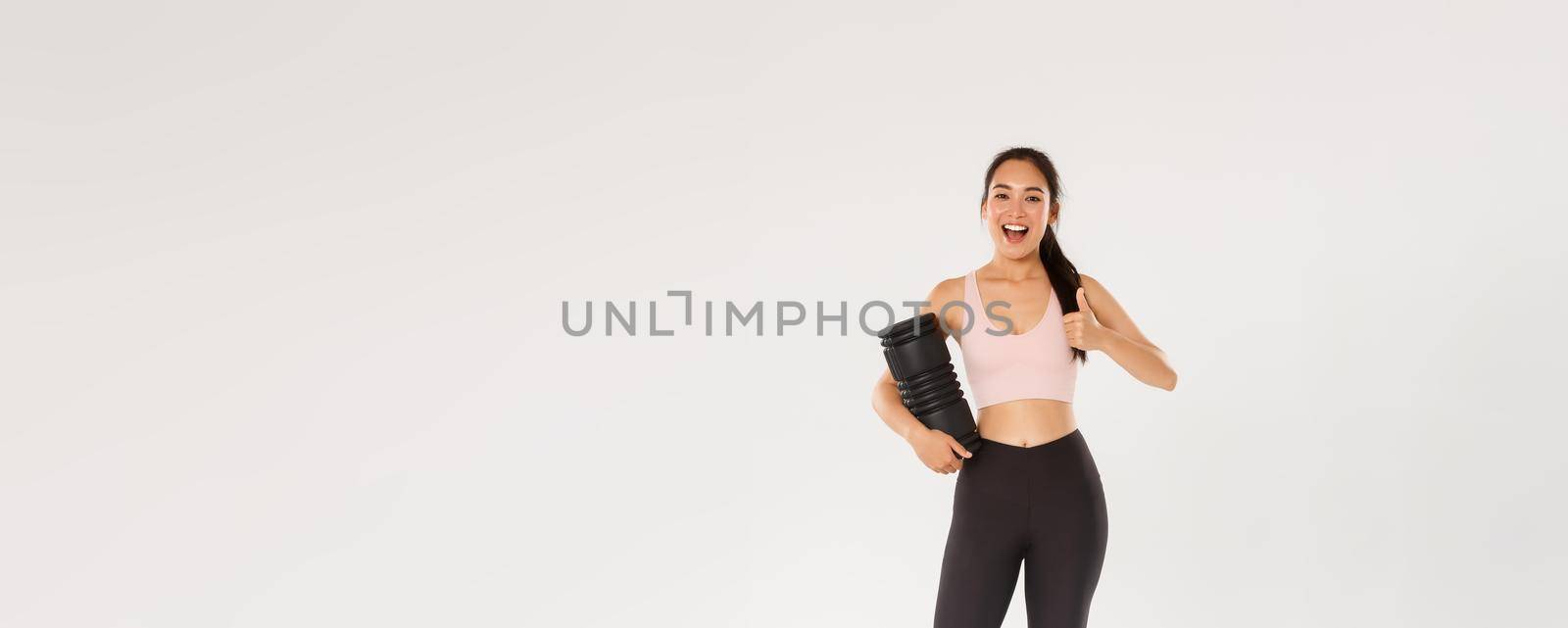 Full length of satisfied smiling asian training coach, fitness girl in sportswear looking pleased, showing thumbs-up, using foam roller after workout to reduce soreness and ache in muscles, recommend by Benzoix