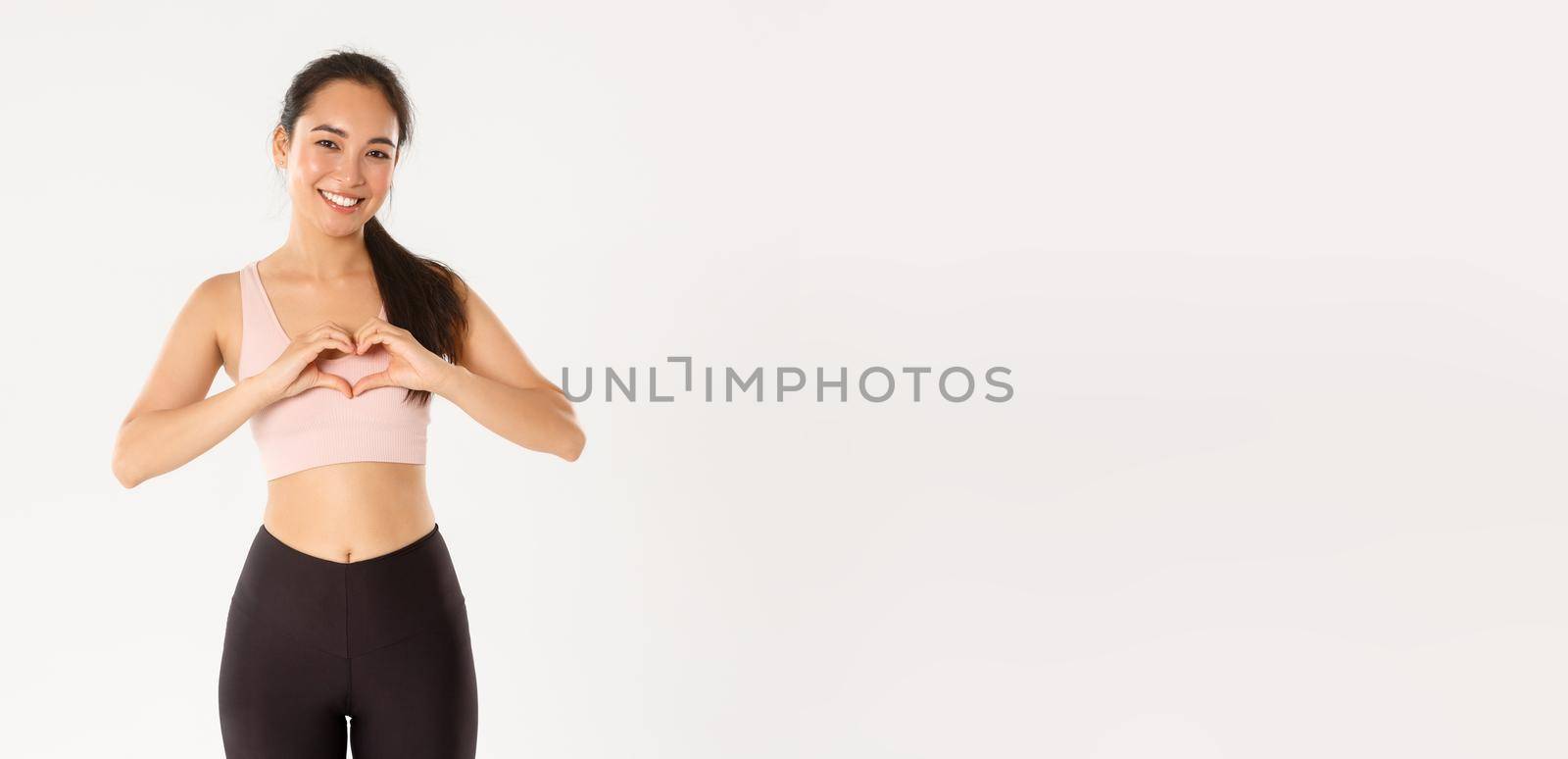 Sport, wellbeing and active lifestyle concept. Smiling asian woman in activewear, sportswoman showing heart gesture, enjoy workout in her favorite gym, using product she loves, white background.