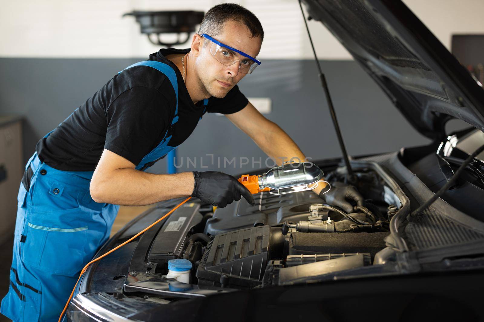Portrait of young caucasian man in blue overalls and safety glasses inspects engine with flashlight. Male car mechanic at work in spacious repair shop. Modern workshop by uflypro