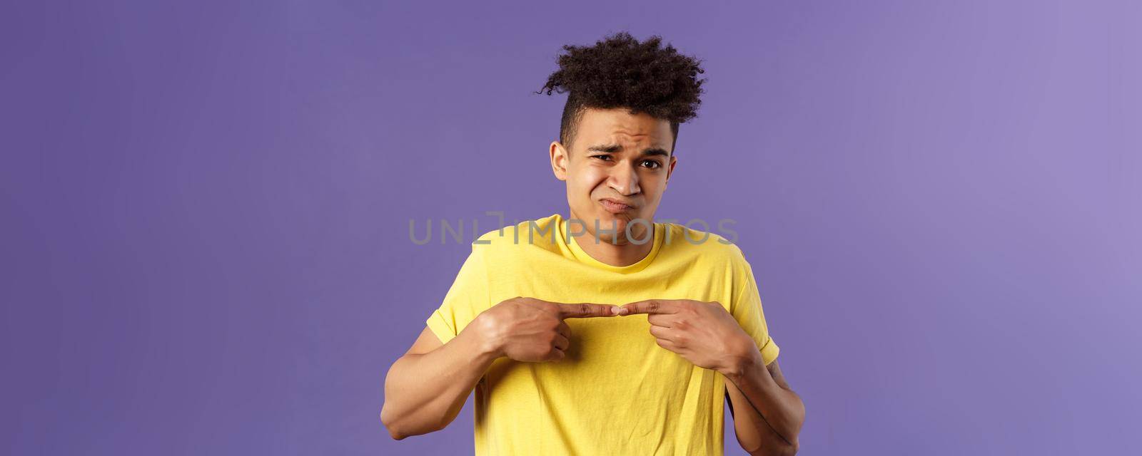 Close-up portrait of shy and modest young silly hispanic man trying say something but being too insecure, grimacing and frowning, look timid, two fingers touching pose, purple background.